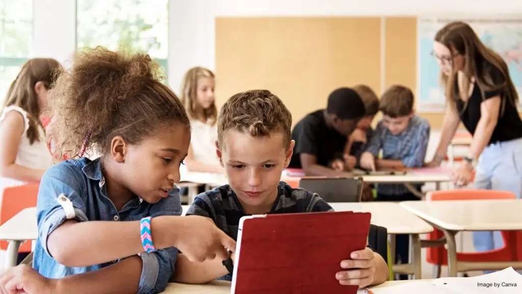Primary students use computer in classroom. The picture shows a girl and a boy shared a laptop, while the teacher and other students in the background.